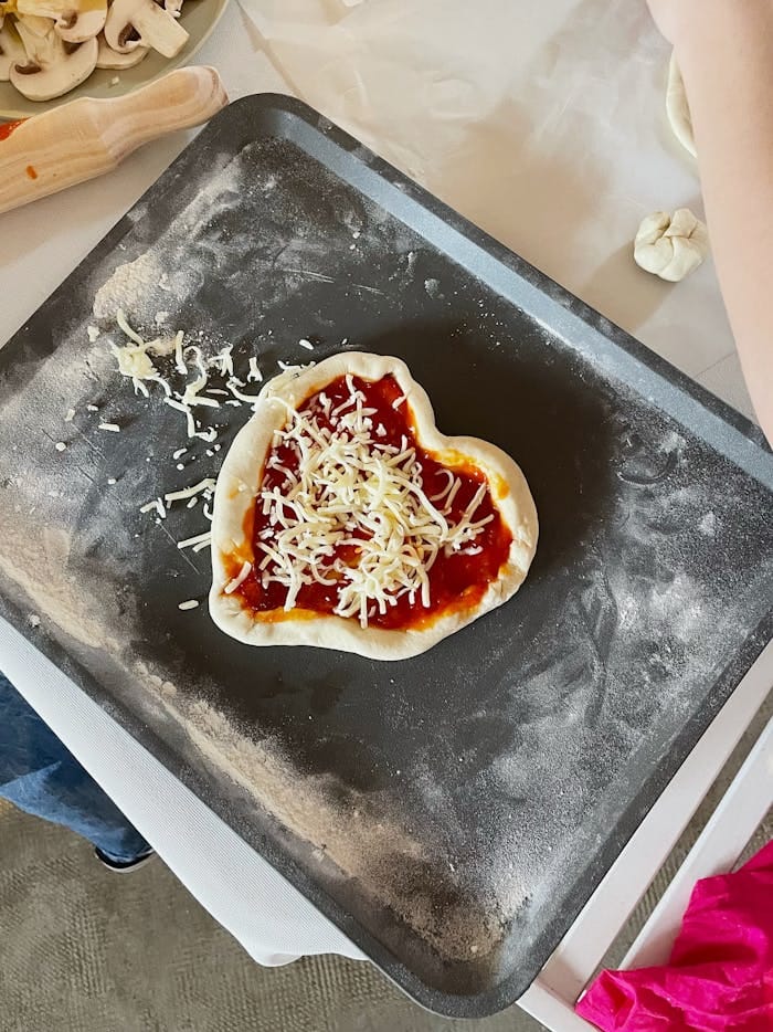 cooking-pizza-in-heart-shape-20204589 Top view of heart-shaped pizza dough with sauce and cheese on a baking tray, ready to bake